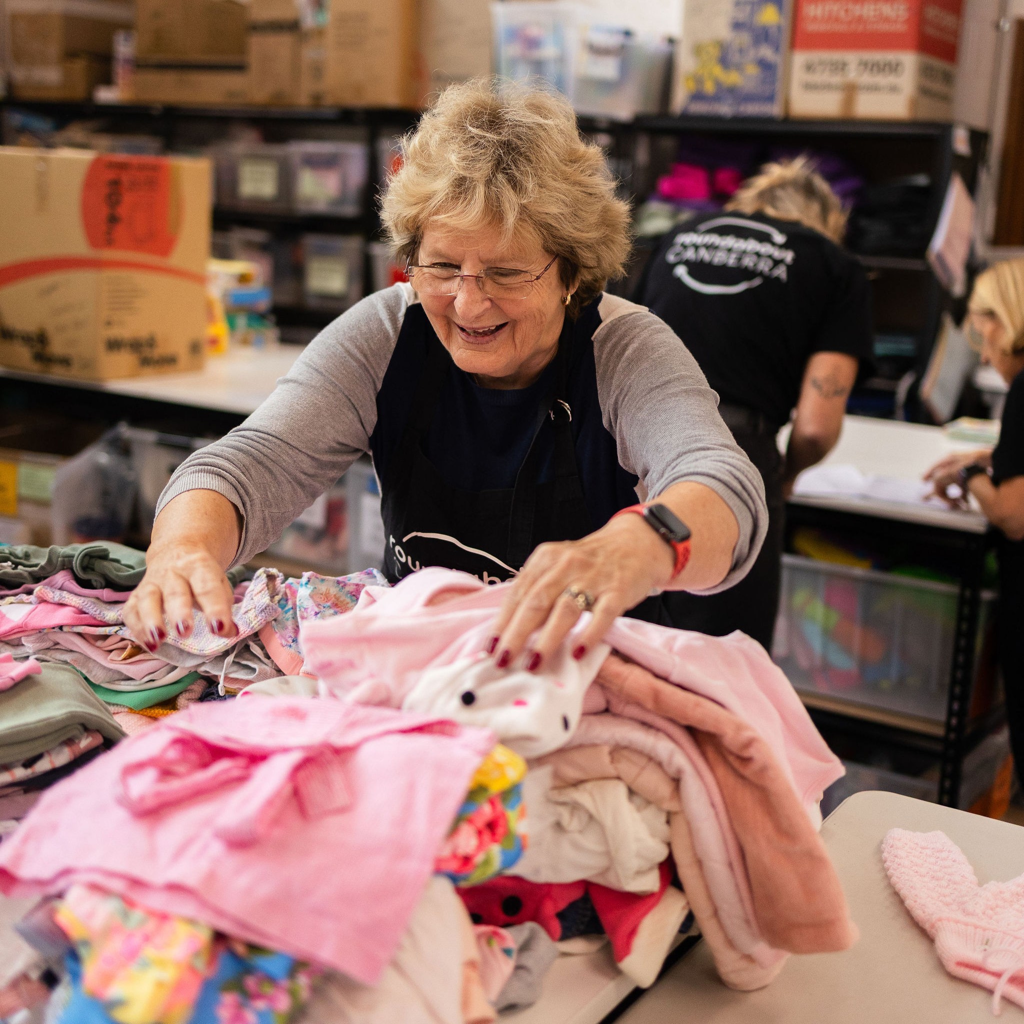 Roundabout Canberra volunteer smiling while sorting piles of donated baby and children’s clothing in a busy donation centre.
