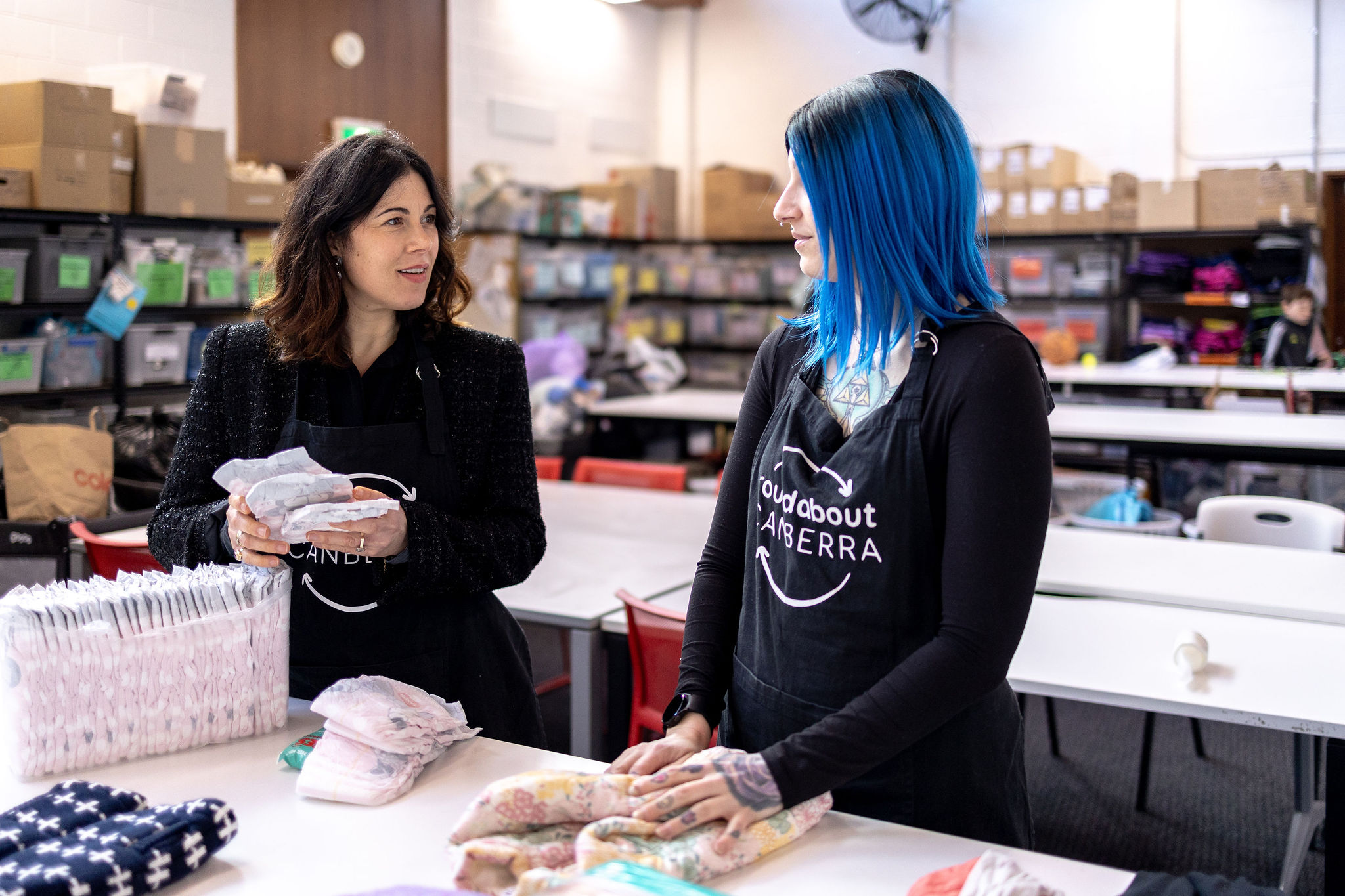 Two Roundabout Canberra volunteers sort and pack donated baby items, including nappies and clothing, while talking in a well-organised donation centre.
