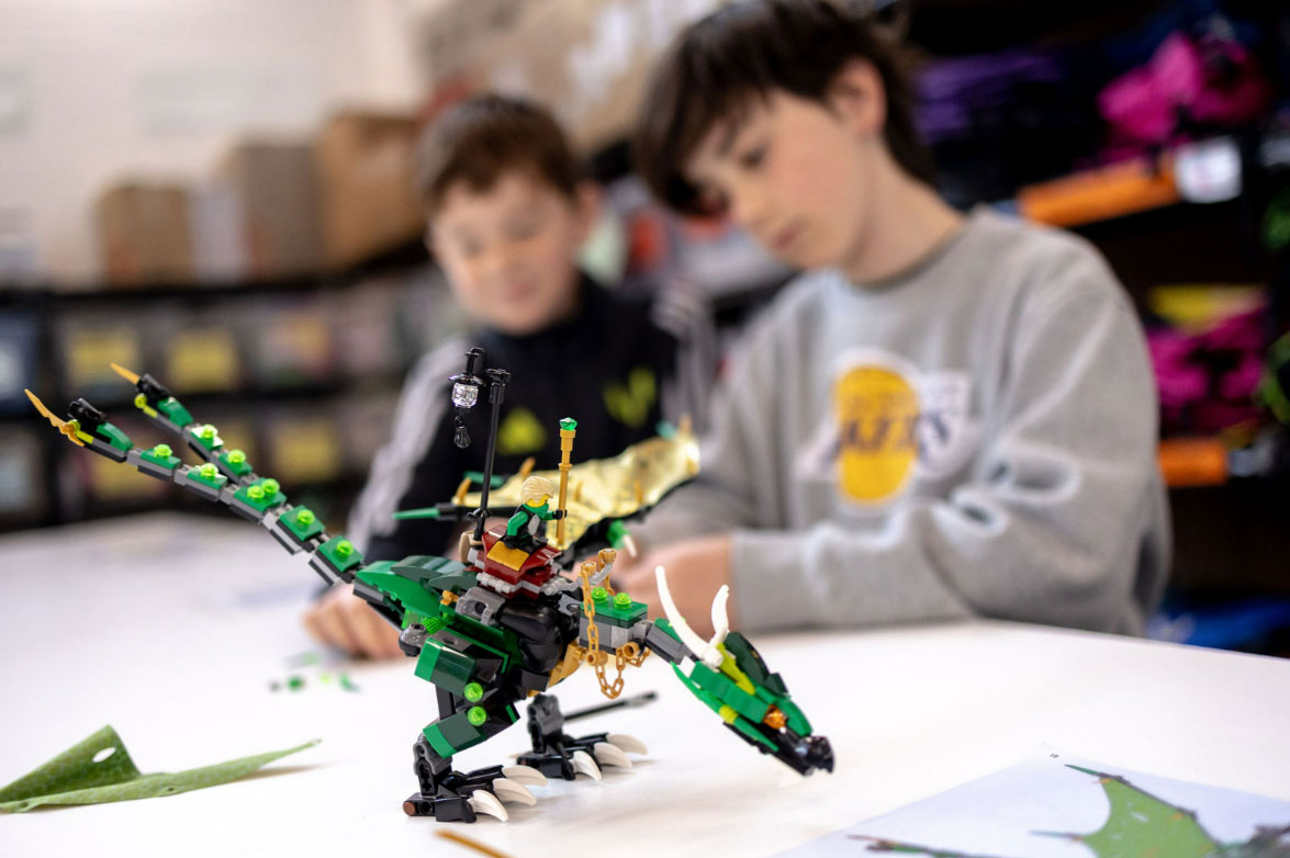 Two children build a green LEGO dragon together at a table, with the detailed model in focus and storage shelves visible in the background.