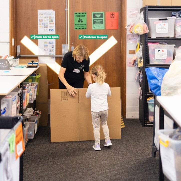 A volunteer helps a young girl hold a cardboard box inside a donation sorting room at Roundabout Canberra, surrounded by shelves of children's items.