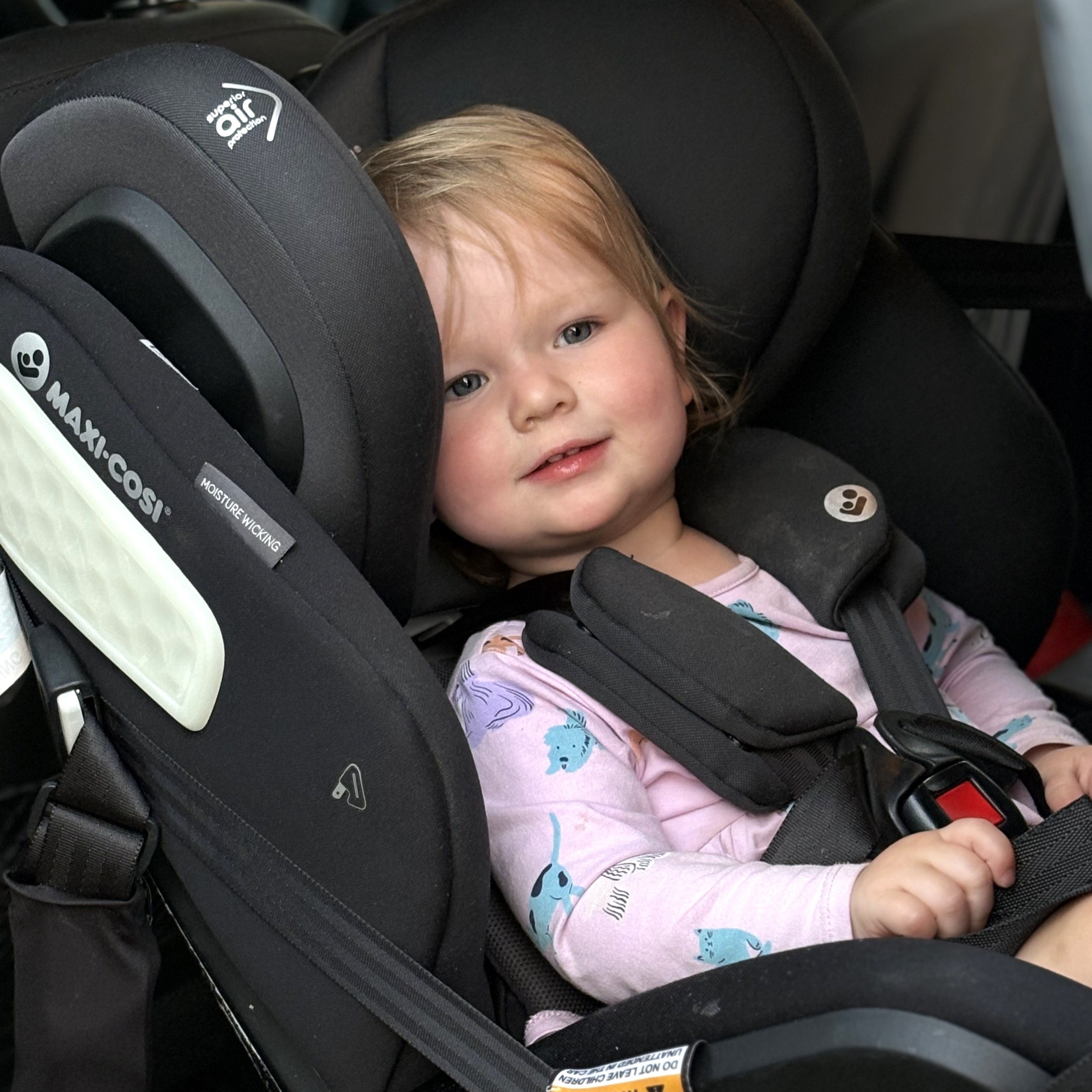 Young child smiling while safely secured in a rear-facing Maxi-Cosi car seat, wearing a pink shirt with animal prints.