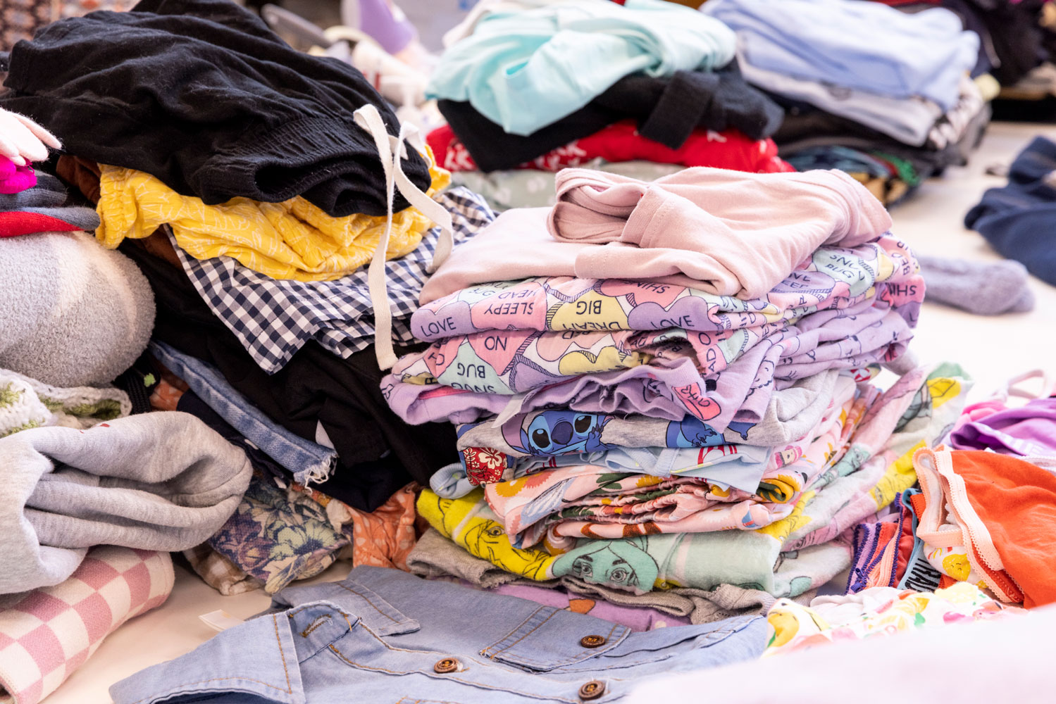 Stacks of folded, preloved children's clothes in a variety of colours and patterns, sorted and ready for distribution at Roundabout Canberra.