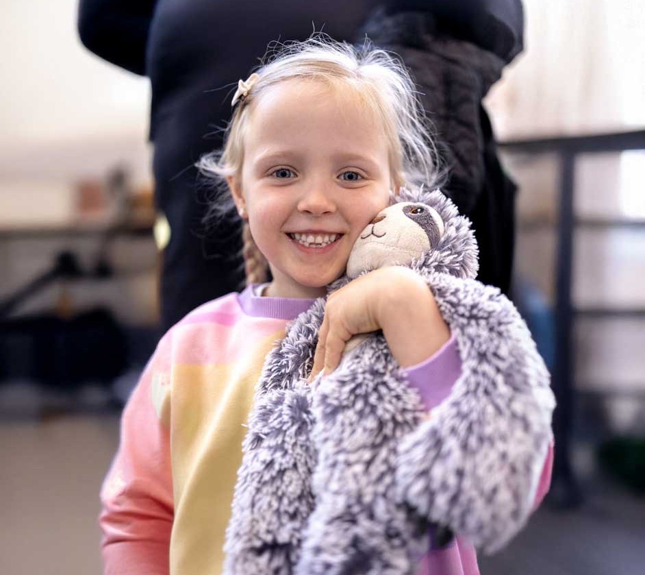 Smiling young girl holding a plush sloth toy close to her face, wearing a colourful sweatshirt at a Roundabout Canberra donation centre.