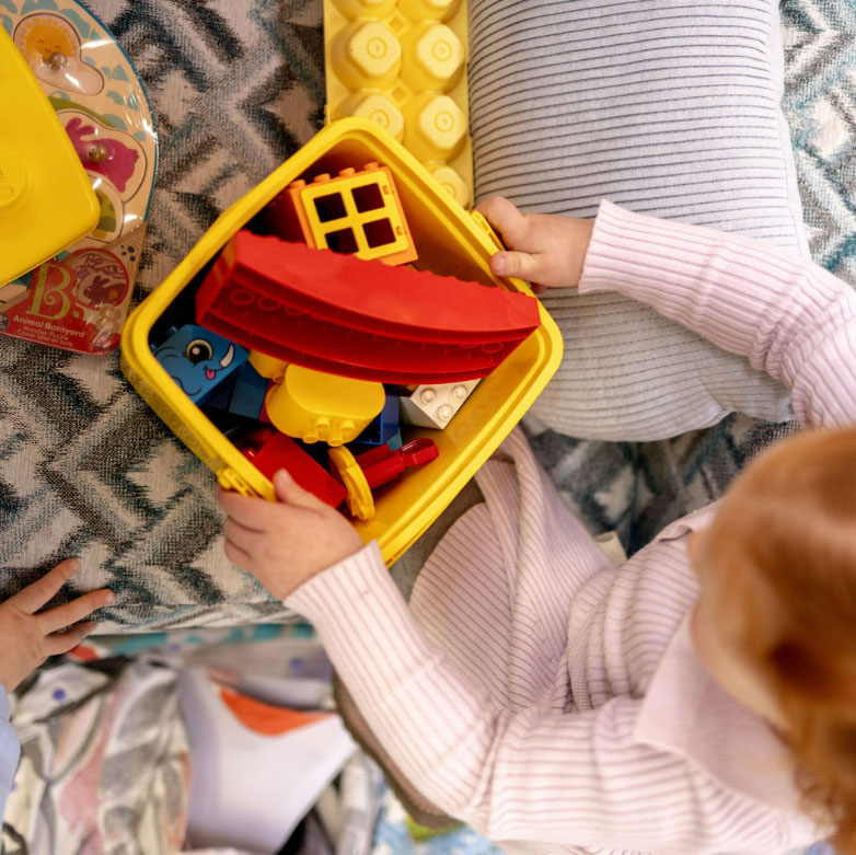 A young child wearing a light pink sweater is holding a yellow plastic bin filled with colorful toy building blocks, including red, yellow, and blue pieces.